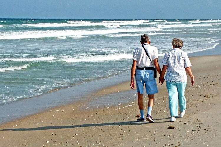 un couple âgé de dos qui marche sur la plage en se donnant la matin