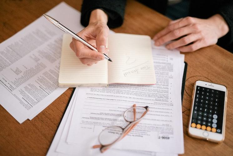 une personne écrit sur son carnet il y'a une calculette et des lunettes sur une feuille