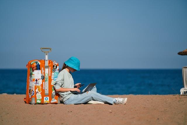 Une femme sur la plage adossée à une valise et son ordinateur portable sur les genoux.