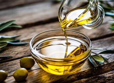 Un filet d'huile d'olive dorée coule d'une carafe dans un bol en verre, posé sur une table en bois rustique, entouré d'olives vertes et de branches d'olivier