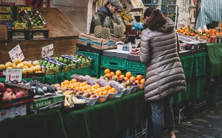 Une femme regarde un stand de fruits et légumes sur un marché. 