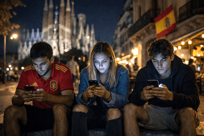 Trois jeunes Espagnols assis de nuit en ville, absorbés par leurs smartphones dont la lumière éclaire leurs visages, avec en arrière-plan une ambiance urbaine animée.