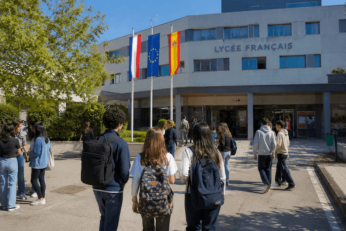 Des élèves se dirigent vers l’entrée d’un lycée français moderne en Espagne, sous les drapeaux français, espagnol et européen. Cette image illustre l’enseignement français à l’étranger et la transition des établissements de la Mission laïque française vers une nouvelle gestion.
