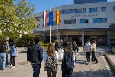 Des élèves se dirigent vers l’entrée d’un lycée français moderne en Espagne, sous les drapeaux français, espagnol et européen. Cette image illustre l’enseignement français à l’étranger et la transition des établissements de la Mission laïque française vers une nouvelle gestion.