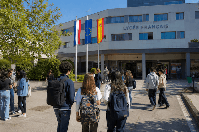 Des élèves se dirigent vers l’entrée d’un lycée français moderne en Espagne, sous les drapeaux français, espagnol et européen. Cette image illustre l’enseignement français à l’étranger et la transition des établissements de la Mission laïque française vers une nouvelle gestion.