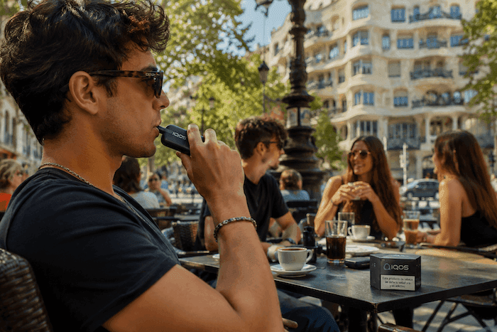 Un jeune adulte utilise un appareil de tabac chauffé IQOS en terrasse d'un café en Espagne, tandis que d'autres clients discutent en arrière-plan, illustrant la popularité croissante de ce produit chez les jeunes adultes.