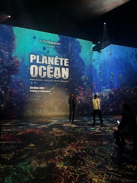 Un homme parle dans un micro dans une grande salle avec une projection derrière lui