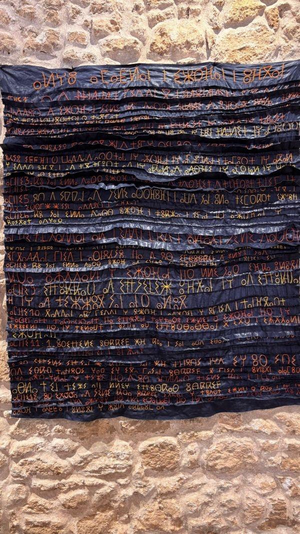 Posée sur le mur de pierres, une « page de bazin africain » teinte en bleu marine porte les signes tifinagh brodés par Soukaïna.  Écrits sur le plissé du tissu, ils reproduisent  une partie de la Déclaration universelle des droits de l'homme.