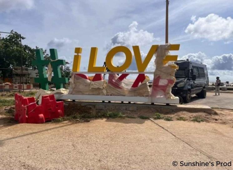 Installation urbaine en cours de montage affichant « I LOVE », partiellement emballée dans du plastique de protection, au bord d’une route.