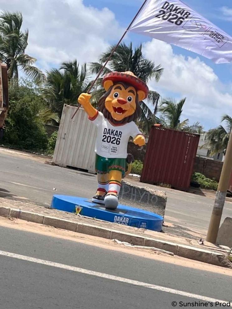 Mascotte des Jeux Olympiques de la Jeunesse Dakar 2026 debout sur un socle bleu, brandissant un drapeau blanc de l’événement, avec des palmiers en arrière-plan.