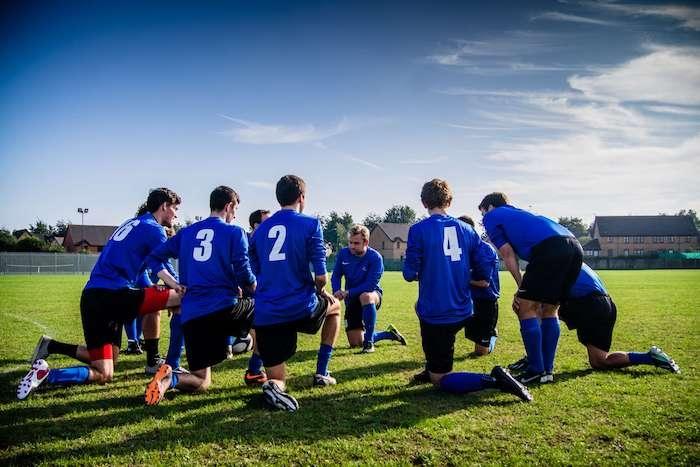 Des jeunes lycéens en maillots de foot en train d'écouter en cercle leur entraineur sur le terrain