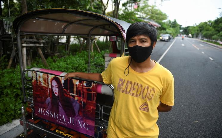 Un chauffeur de tuk-tuk devant son vehicule et une affiche de K-pop