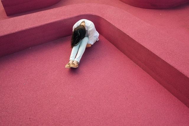 Une jeune femme assise seule dans un espace aux tons roses vifs, la tête baissée, ses bras entourant ses jambes