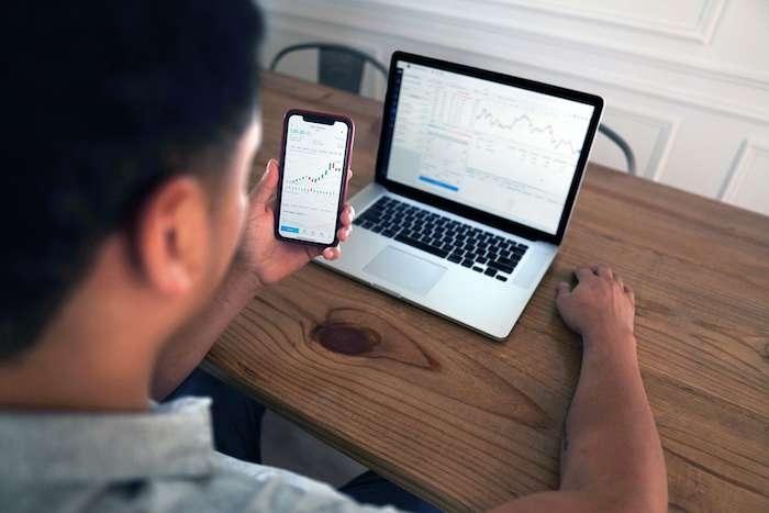 un homme en train d'investir devant son ordinateur et de regarder les cours la bourse sur son téléphone