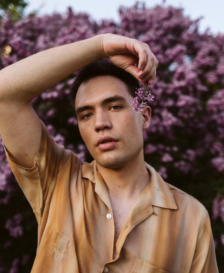 Un homme pose, une fleur à la main et un arbre en fleurs roses derrière lui