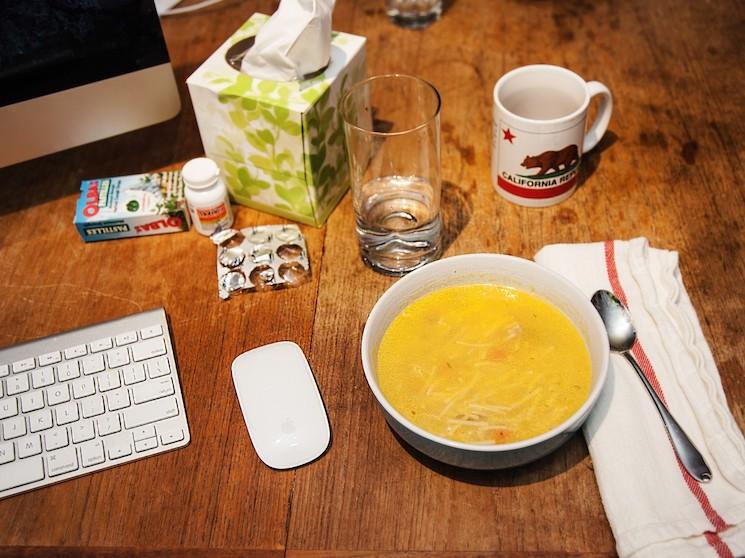 Personne travaillant à son ordinateur tout en étant malade (soupe, mouchoirs, tasse)