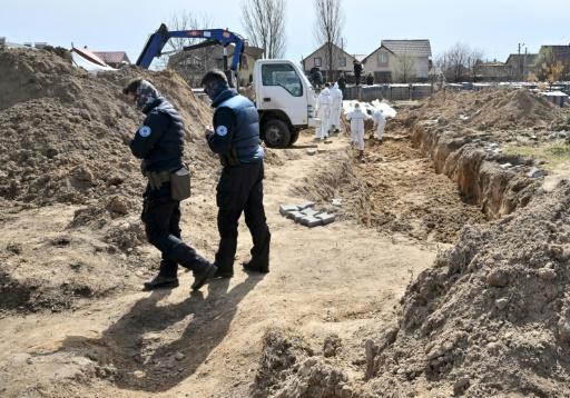Un gendarme français, accroupi, fait vrombir dans le ciel de Boutcha un drone photographique. A ses pieds, la fosse commune s'étend sur 14 mètres dans la terre sableuse