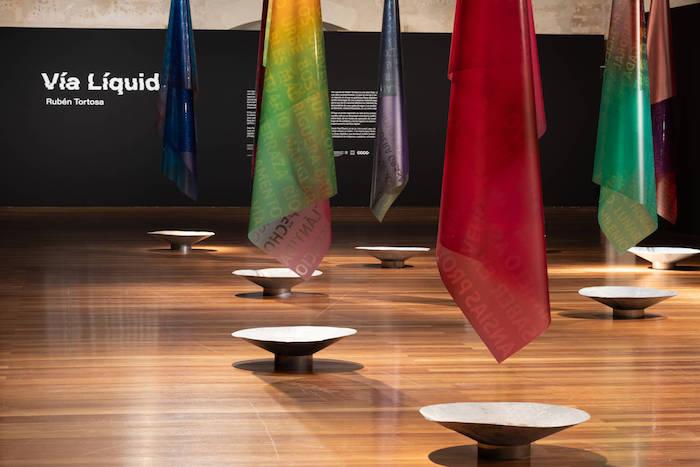 des drapeux de plusieurs couleurs suspendus à l'envers dans l'expo Rubén Tortosa.Vía Líquida