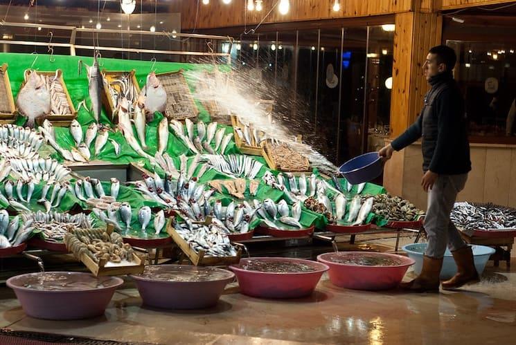 Marché de poissons à Kumkapı, Istanbul, avec des étals colorés et un poissonnier arrosant les produits pour les garder frais.