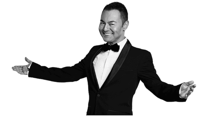 Portrait de Serdar Ortaç, icône de la pop turque, vêtu d’un élégant costume noir, souriant et accueillant, promouvant son concert à Izmir en janvier 2025.