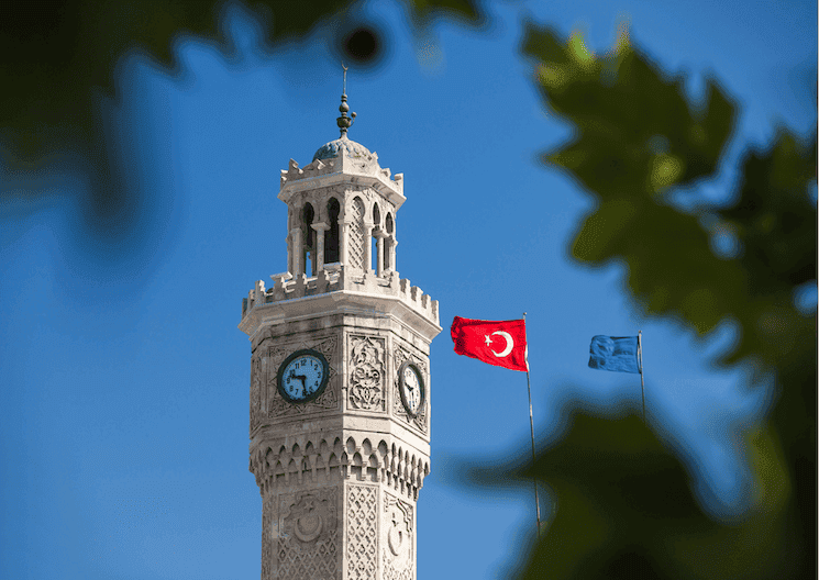 Photo de la Tour de l’Horloge d’Izmir en janvier, entourée d’un ciel bleu éclatant et de drapeaux flottants, un monument emblématique au cœur de la place Konak.