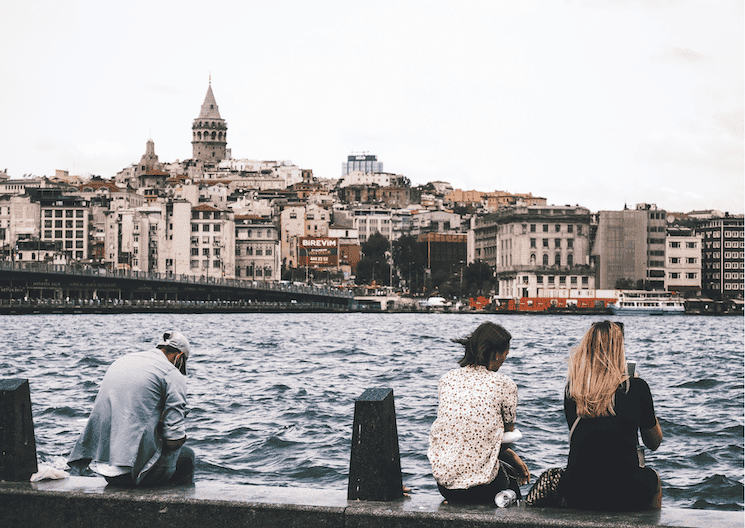 Trois jeunes assis au bord du Bosphore à Istanbul, absorbés par leurs téléphones, illustrant la FOMO et l’isolement numérique.