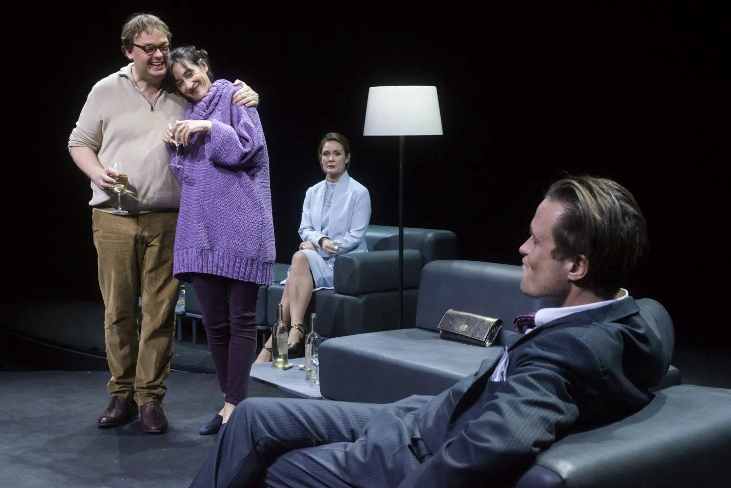 Un homme de dos assis dans un fauteuil fait face à un homme et une femme enlacés et debout. Derrière eux, une femme est assiste et les regarde