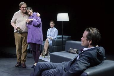 Un homme de dos assis dans un fauteuil fait face à un homme et une femme enlacés et debout. Derrière eux, une femme est assiste et les regarde