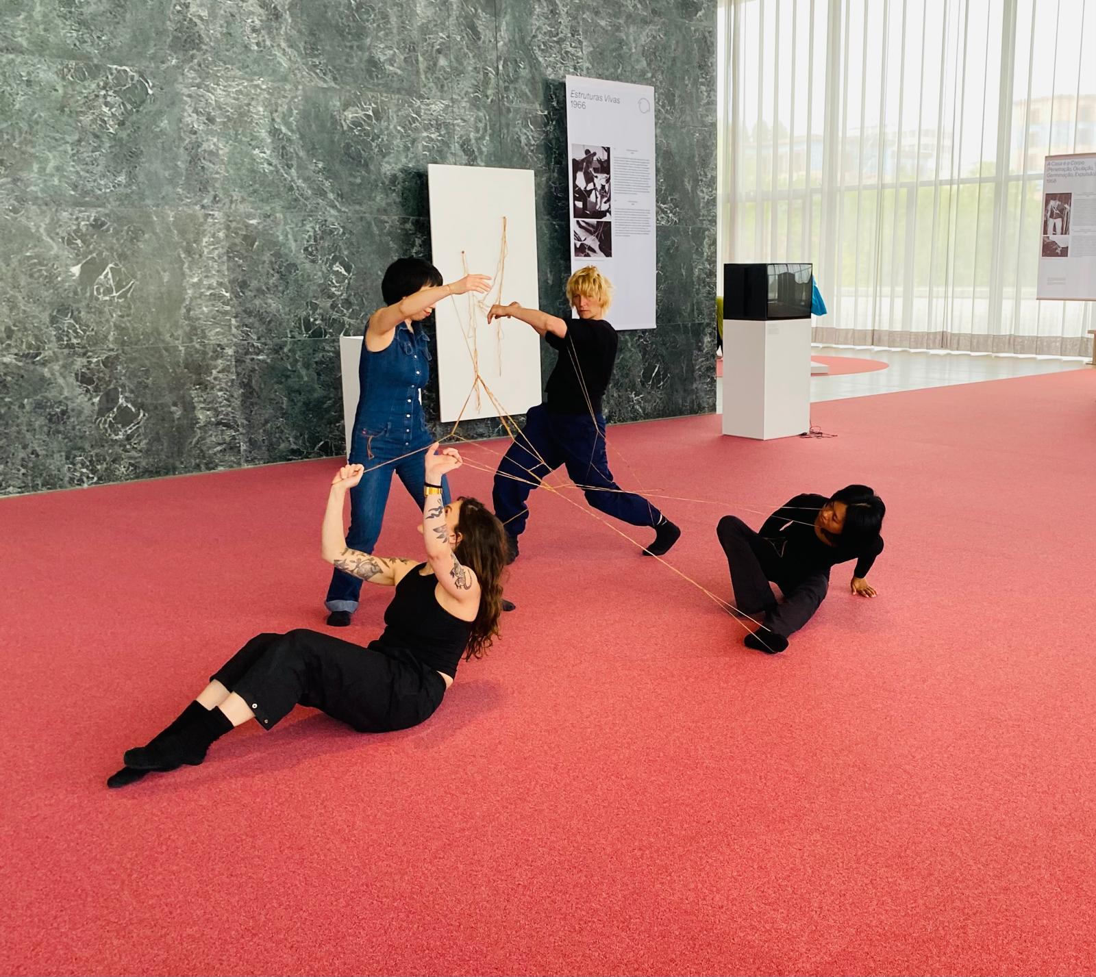 Quatre femmes dansent en étant reliées par des élastiques, reproduisant la "Performance avec fils" de Lygia Clark