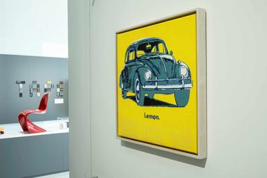 Vue de l'exposition, un tableau au fond jaune avec une voiture bleue est accroché à un mur, des objets designs sont présents en arrière plan