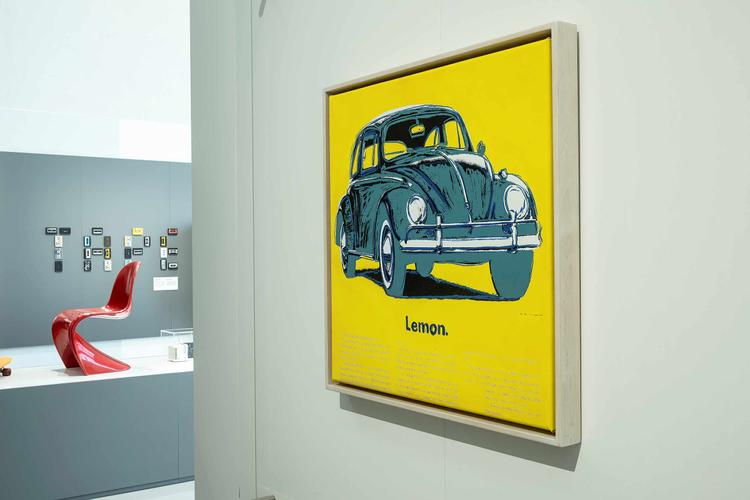 Vue de l'exposition, un tableau au fond jaune avec une voiture bleue est accroché à un mur, des objets designs sont présents en arrière plan