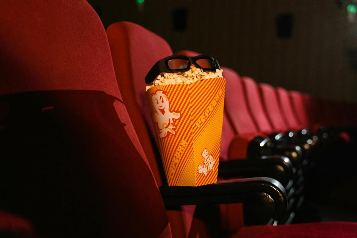 Lunette de cinéma posées sur un paquet de pop-corn dans une salle de cinéma. Le paquet est déposé sur l'accoudoir d'un siège rouge de cinéma.