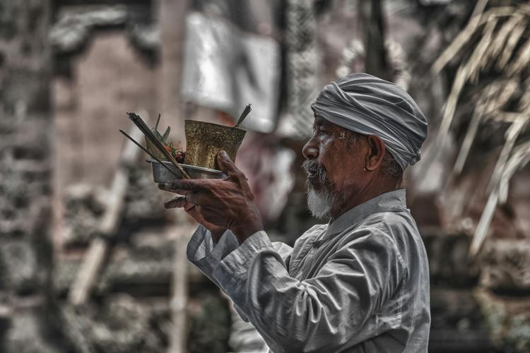 un homme en turban qui tient un bol métallique avec des offrandes et de l'encens devant un temple