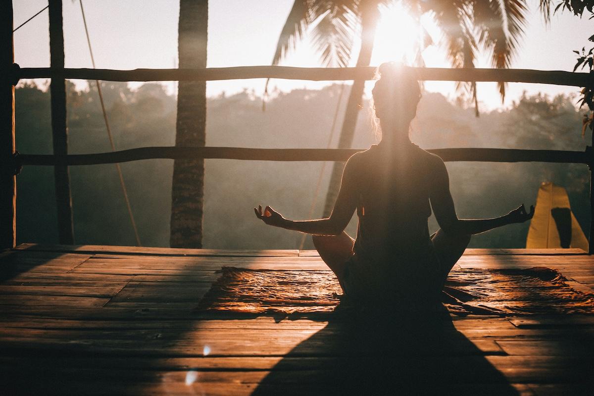 une femme assise en posture de yoga de dos avec un palmier en fond