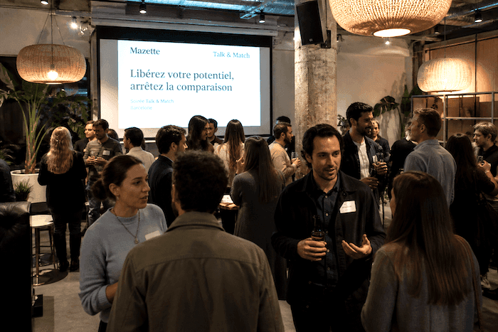 Participants échangeant lors d’un événement de networking à Barcelone, dans un espace moderne et chaleureux, avec un écran affichant “Libérez votre potentiel, arrêtez la comparaison”.