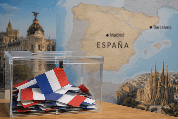 Une urne électorale transparente remplie de bulletins aux couleurs françaises est posée au premier plan. En arrière-plan apparaissent une carte de l’Espagne ainsi que des vues emblématiques de Madrid et Barcelone, illustrant les élections consulaires des Français établis en Espagne.