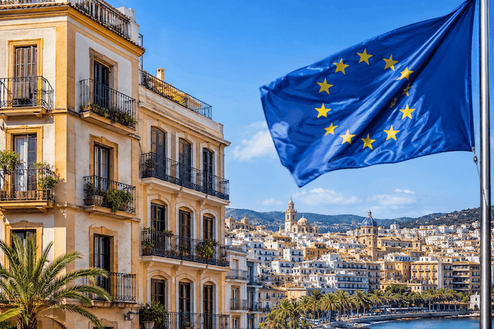 Un immeuble résidentiel de style méditerranéen surplombe une ville côtière espagnole baignée de soleil, tandis qu'un drapeau de l'Union européenne flotte au premier plan. Cette illustration évoque les enjeux fiscaux et immobiliers liés aux investissements des non-résidents en Espagne.