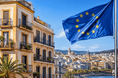 Un immeuble résidentiel de style méditerranéen surplombe une ville côtière espagnole baignée de soleil, tandis qu'un drapeau de l'Union européenne flotte au premier plan. Cette illustration évoque les enjeux fiscaux et immobiliers liés aux investissements des non-résidents en Espagne.