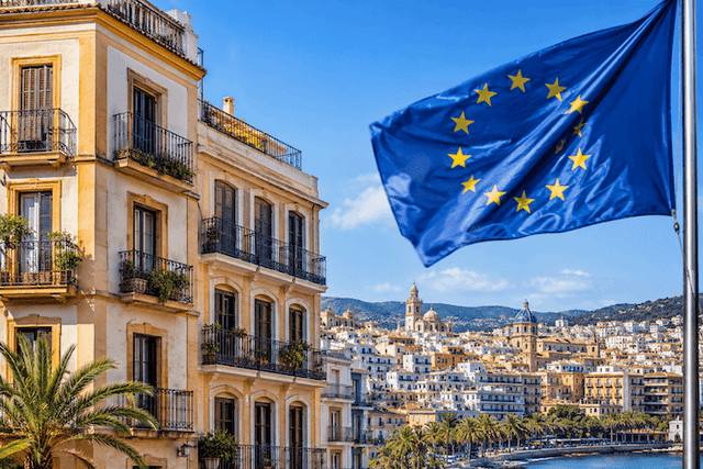 Un immeuble résidentiel de style méditerranéen surplombe une ville côtière espagnole baignée de soleil, tandis qu'un drapeau de l'Union européenne flotte au premier plan. Cette illustration évoque les enjeux fiscaux et immobiliers liés aux investissements des non-résidents en Espagne.