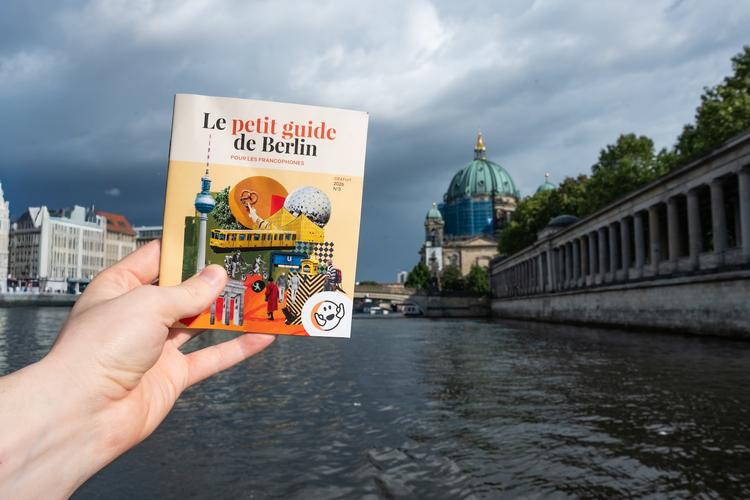 Une main tenant le petit guide de Berlin 2026 avec le Berliner Dome en fond d'image.