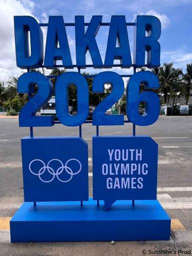 Panneau bleu « DAKAR 2026 » installé en extérieur, avec les anneaux olympiques et l’inscription « Youth Olympic Games » au premier plan.