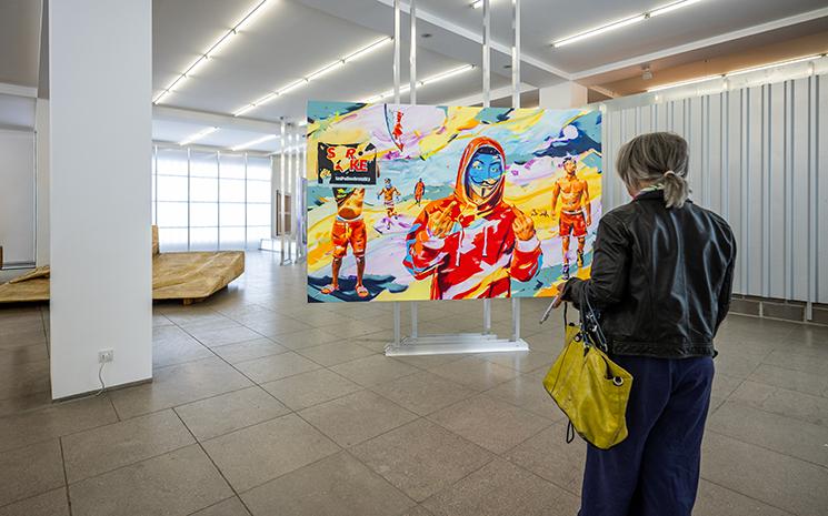 Une personne observe une toile très colorée de l'artiste Bisky représentant des scènes de révoltes