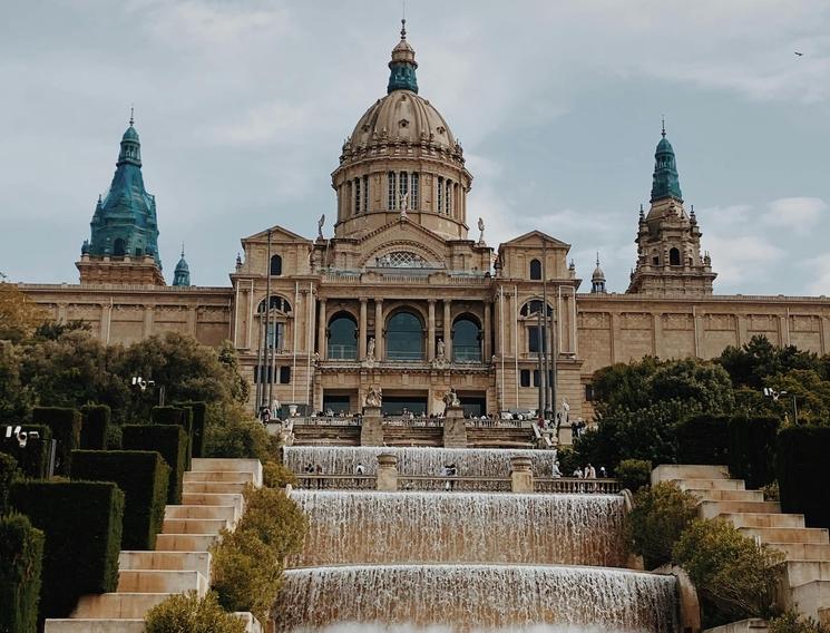 Vue en contre-plongée du Palais National de Montjuïc, monument emblématique de l'architecture barcelonaise. Au premier plan, des cascades d'eau symétriques dévalent les paliers, encadrées par d'imposants escaliers de pierre et des haies taillées. L'édifice, surmonté de son dôme central et de ses tours aux toits verts, surplombe l'esplanade où déambulent quelques passants sous un ciel couvert