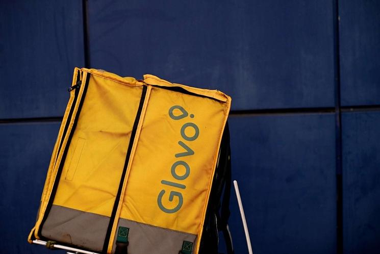 Sac jaune de livreur, avec une inscription verte "Glovo", posé sur le sol sur un fond bleu.