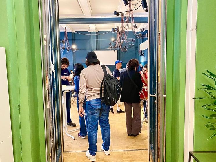 Entrée de la salle, plusieurs personnes entrent, un bénévole près d'une table haute donne des autocollants avec les prénoms de chacun