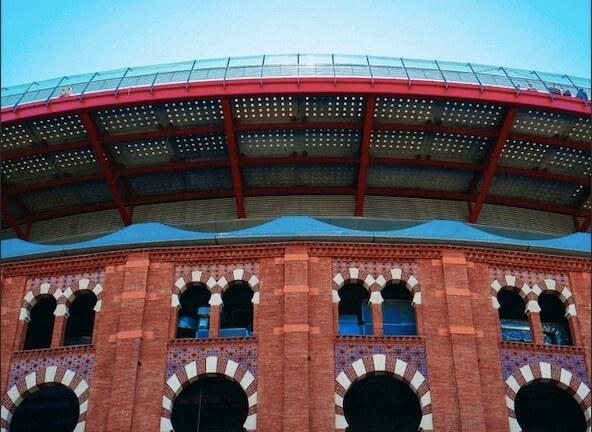 Vue en contre-plongée de la façade en briques des anciennes arènes de Barcelone, reconverties en centre commercial (Las Arenas). La structure historique est surmontée d'un rooftop moderne avec un toit circulaire offrant une vue sur Barcelone. Des visiteurs sont visibles sur la plateforme d'observation sous un ciel bleu