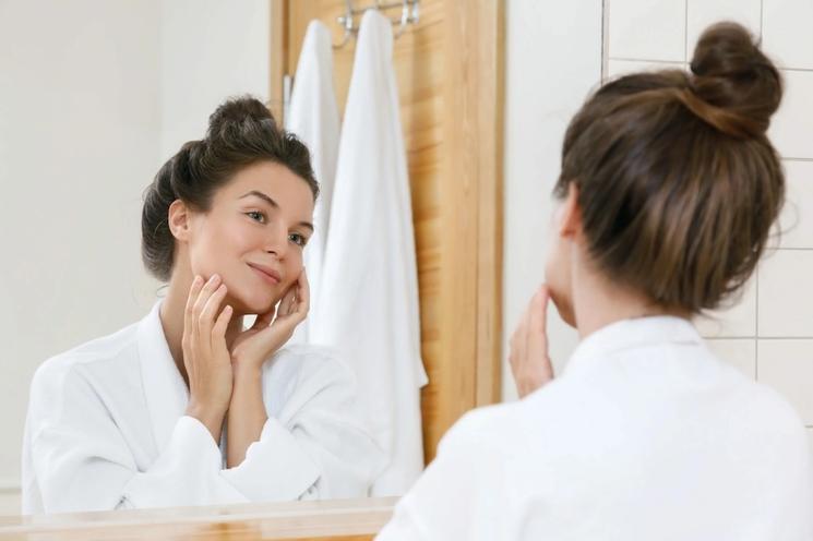 femme avec un chignon qui se regarde dans le miroir de sa salle de bain blanche en peignoir blanc