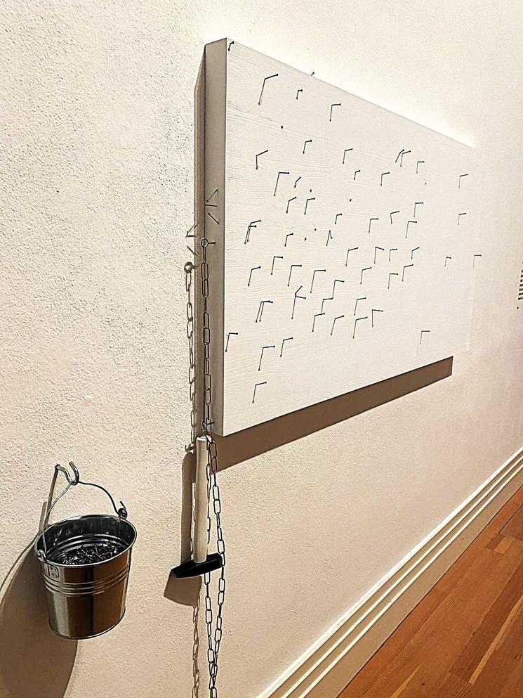 Toile blanche avec clous plantés dedans. A côté, un marteau et un sceau rempli de clous