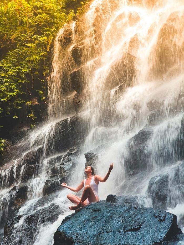une femme en posture de méditation sur un rocher avec une cascade derrière elle qui s'écoule sur une falaise en escalier