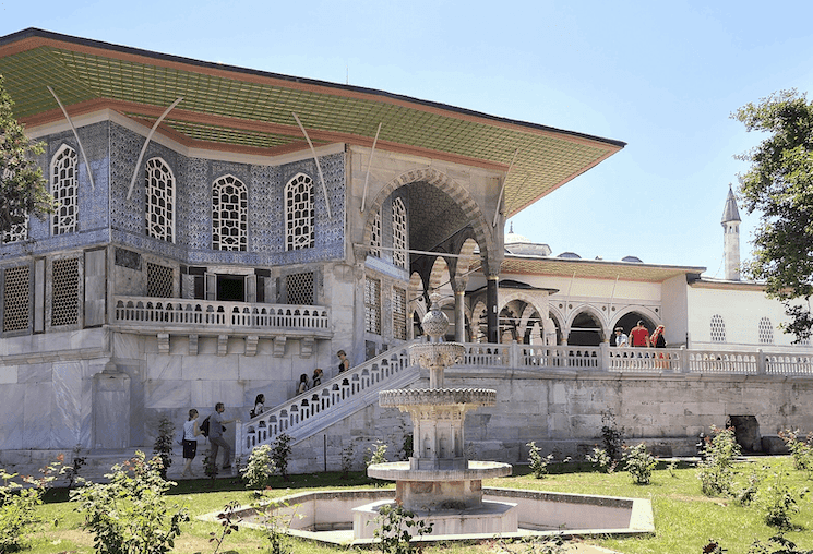 Vue du Palais de Topkapı à Istanbul, mettant en avant une façade ornée de mosaïques, des arches élégantes et une fontaine au premier plan sous un ciel bleu.